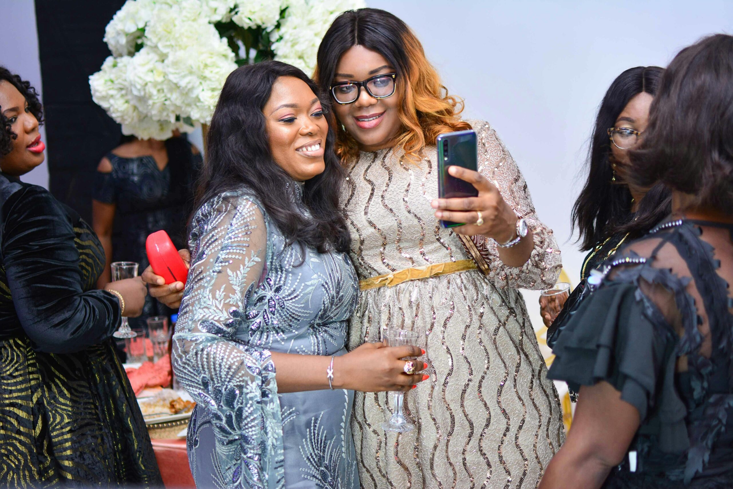 Two black women taking a selfie at a Happy New Year's party. One has on a blue sequin dress and the other one has on a silver dress.